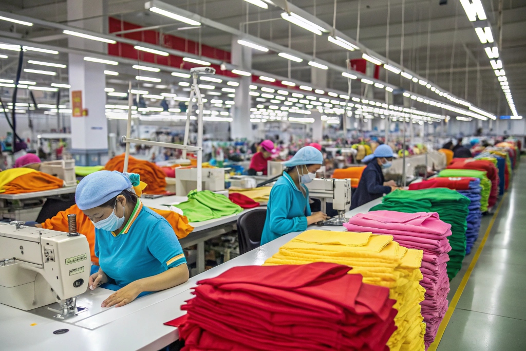 Factory workers sewing garments surrounded by colorful stacks of fabric on tables.