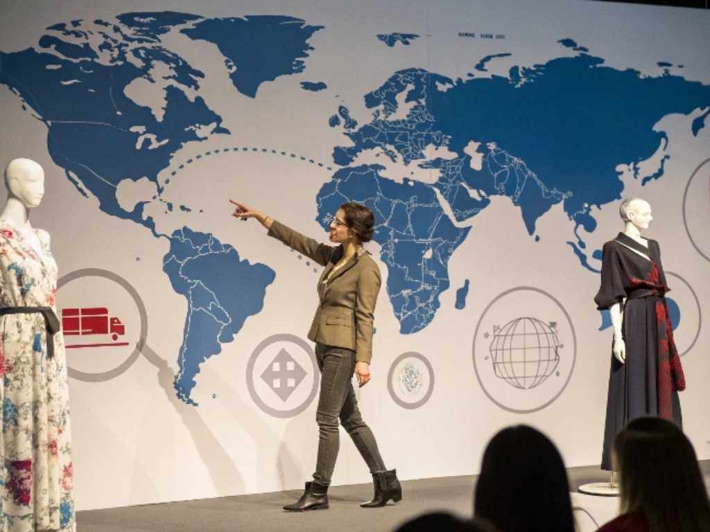 Fashion presenter points at world map backdrop with mannequins on stage.